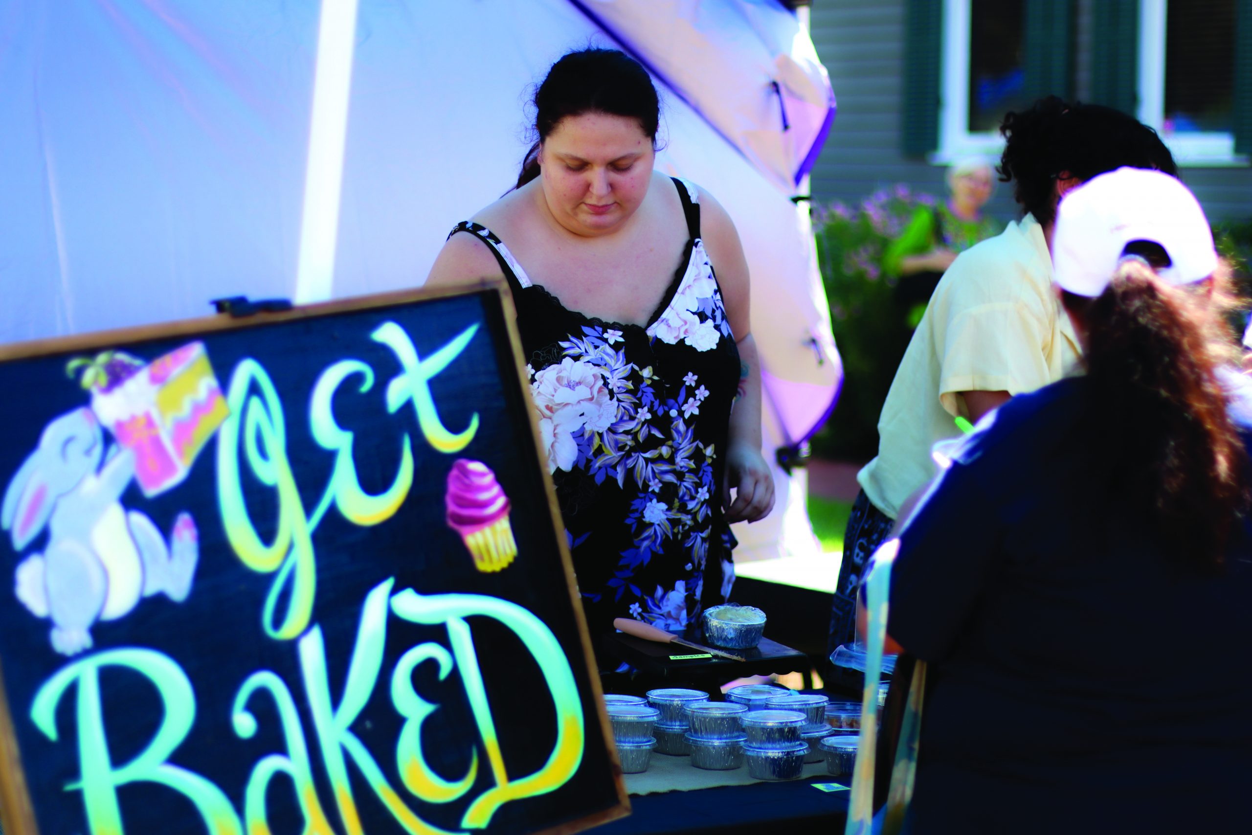 A bakery worker tends to a table of small, cup shaped baked goods. A large sign with the name "Get Baked" in the foreground.